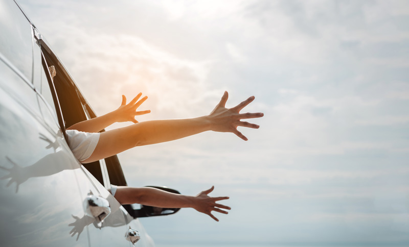 Passengers stretching their arms out of a car window, enjoying a carefree drive under a sunny sky